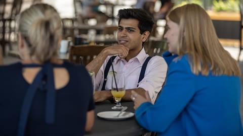 A person in a pink shirt sits at a table with two others, engaged in conversation. They have a tropical drink in front of them.