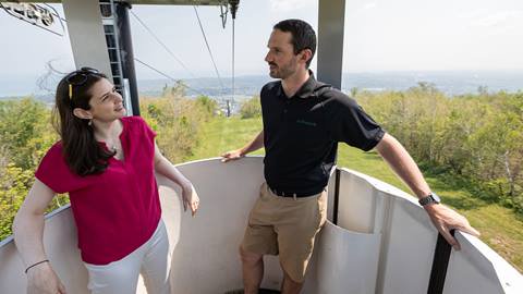 Two people standing inside a gondola lift with an outdoor scene featuring trees, grass, and a distant city view in the background.