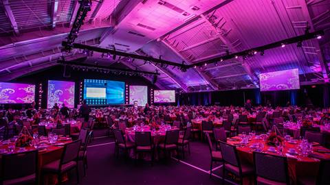 A large event hall with purple lighting is set up with round tables and chairs, ready for a banquet. Screens and decorations are visible on the stage at the far end.