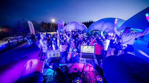 A DJ performs outdoors at night in front of a lively crowd under tents, illuminated by colorful lights.