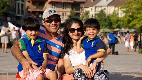 A family of four smiling outdoors on a sunny day, with two children sitting on their parents' laps.