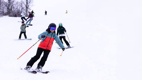 A group of people skiing down a snowy slope, with one person in the foreground wearing an orange and blue jacket and black helmet.