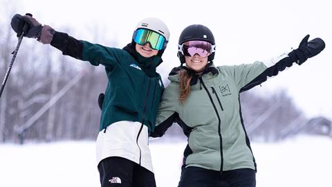 Two skiers in goggles and helmets pose on a snowy slope, smiling and raising their arms.