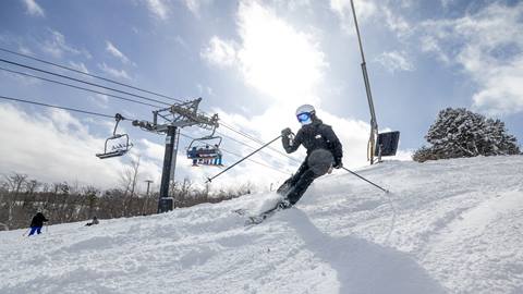 A skier descends a snowy slope under a bright sky, with a ski lift and seated skiers in the background. Snow-covered trees are visible on the right.
