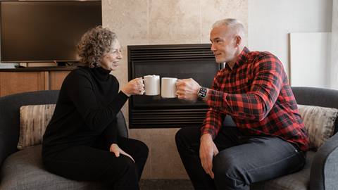 Two people sitting across from each other on a couch, smiling and clinking white mugs, in front of a fireplace.