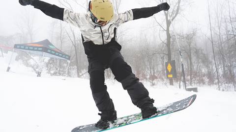 A person wearing winter gear is snowboarding down a snowy hill. Trees and a tent are visible in the background on a foggy day.