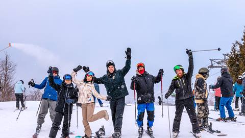 A group of people in winter clothing and ski gear stand on a snowy slope, posing with arms raised and smiling. Other skiers and snowboarders can be seen in the background.