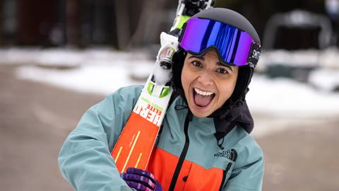 Person in a green and orange jacket, black helmet, and purple ski goggles smiles while holding skis over their shoulder in a snowy environment.