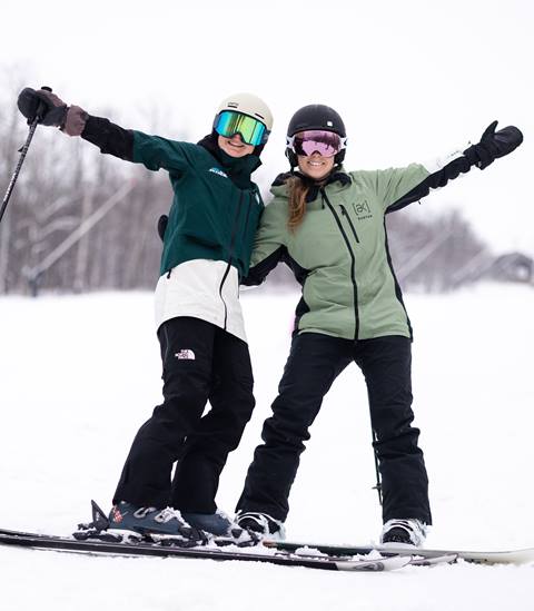 Two skiers in goggles and helmets pose on a snowy slope, smiling and raising their arms.