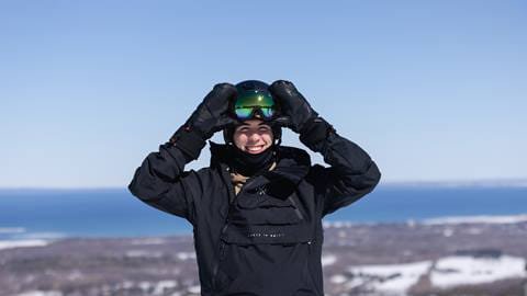 A person wearing black ski gear and a helmet stands on a snowy hill, smiling. The background shows a clear sky and a distant body of water.