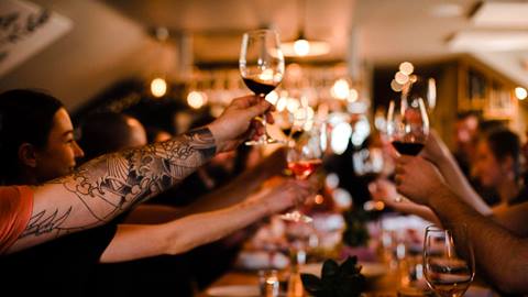 People toasting with red wine at a bustling, dimly lit restaurant.