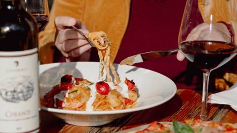 A person eating shrimp pasta at a restaurant table, with a glass and bottle of red wine nearby.