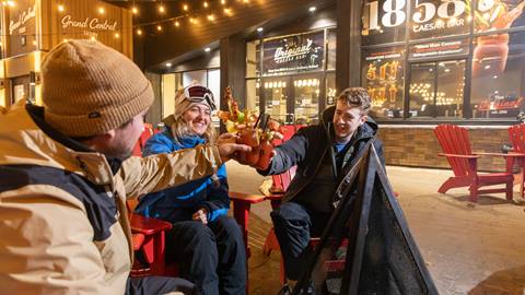 Three people in winter clothing toast drinks while sitting on outdoor red chairs at night. Festive lights and restaurant signage are in the background.