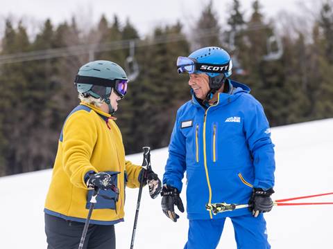 Two people in ski gear stand on a snowy slope, conversing. One wears a yellow jacket, the other a blue one. Ski poles are in their hands, and trees are in the background.