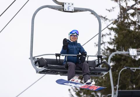 Person in blue jacket and goggles sitting on a ski lift, holding a snowboard, with snowy trees in the background.