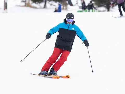 A person in a blue and black jacket and red pants skis downhill on a snowy slope.