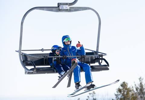 Two skiers wearing blue outfits and goggles ride a ski lift on a clear day, surrounded by trees.