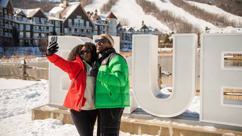 Two people in colorful winter jackets take a selfie in front of large letters spelling "BLUE" at a snowy resort with buildings and a ski slope in the background.