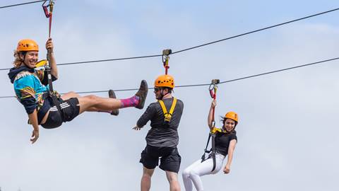 Three people wearing helmets and harnesses ride on zip lines in the sky against a cloudy background.