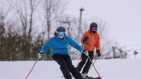 Two people skiing on a snowy slope, wearing helmets and goggles. One skier in a blue jacket is in the foreground, the other in an orange jacket is behind.