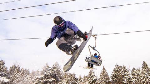 Snowboarder performing a mid-air trick against a snowy forest backdrop, with a ski lift and skiers visible nearby, and a pink gradient on the left.