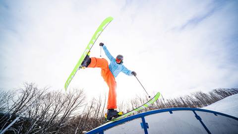 Skier in bright gear performs a trick on a rail, with one leg raised, against a backdrop of bare trees and cloudy sky.