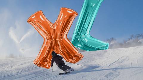 Skier descends a snowy slope, turning sharply around large inflatable balloons shaped like the letters "X" and "L" under a clear blue sky.