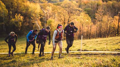 A group of people in athletic gear hike uphill on a grassy trail surrounded by autumnal trees.