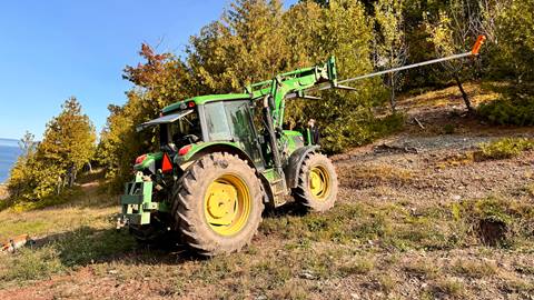 A green tractor with large wheels is parked on a grassy slope. It has an extended arm pointing outward. Trees with green and some brown leaves surround the area.